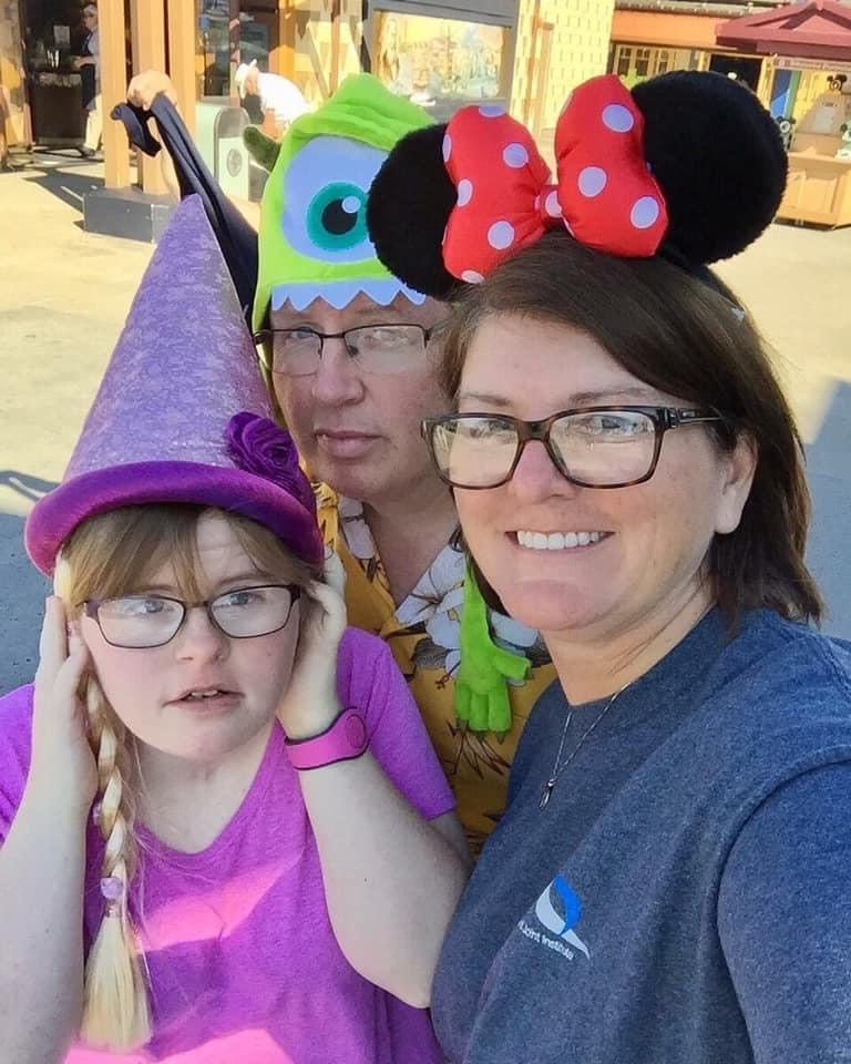 On the left is Emily, a young woman with Down syndrome, in a pink princess cap. In the middle is Tom, a middle aged white man, with a Monster's Inc. hat on. On the right is Tina, a middle aged white woman, wearing a Minnie Mouse headband. They are all smiling at the camera, Emily is covering her ears.