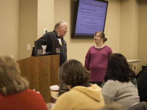 On left, is middle-aged white man Tom Felter talking to Emily Felter, his 26 year old daughter who has Down syndrome. They are presenting in front of a crowd. Three women are visible from the back in the front row, and there is a TV screen presenting a powerpoint slide.