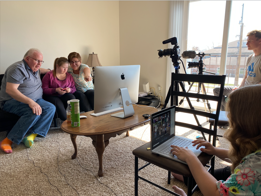 Picture of Tom, Emily, and Tina sitting in front of an iMac answering questions on a Zoom call. Tina is pointing to the screen, and Tom and Emily are looking intently at it. To the right, there is a blonde-haired woman managing the Zoom meeting on her laptop, and a young man operating two cameras pointed at Tom, Tina, and Emily.